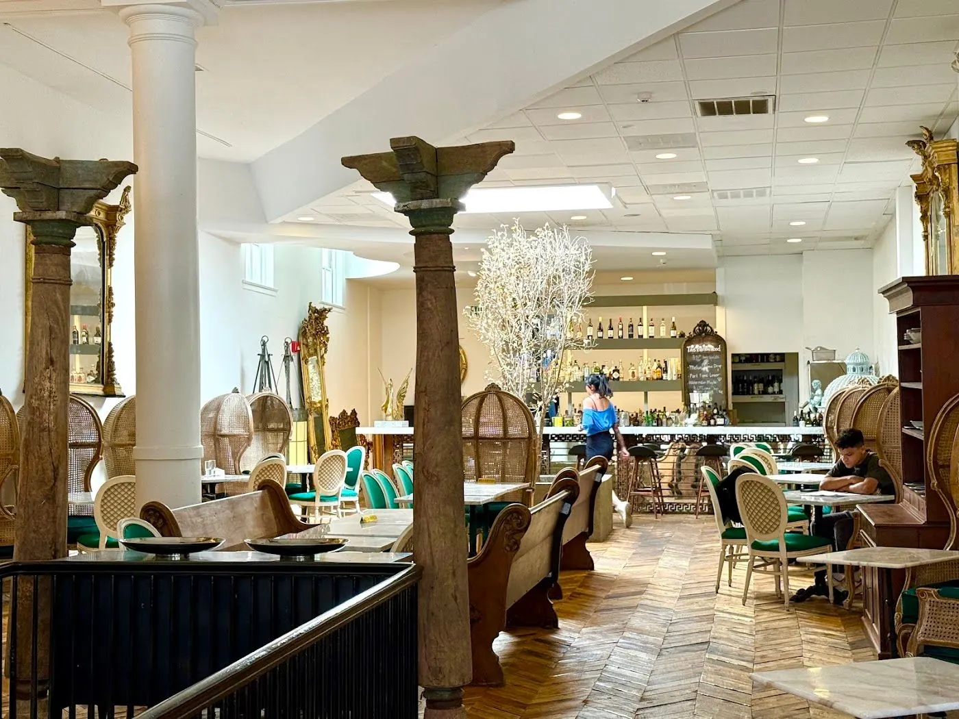 The long dining room: carved wooden columns, marble tables, green velvet chairs and the gold tree under the skylight