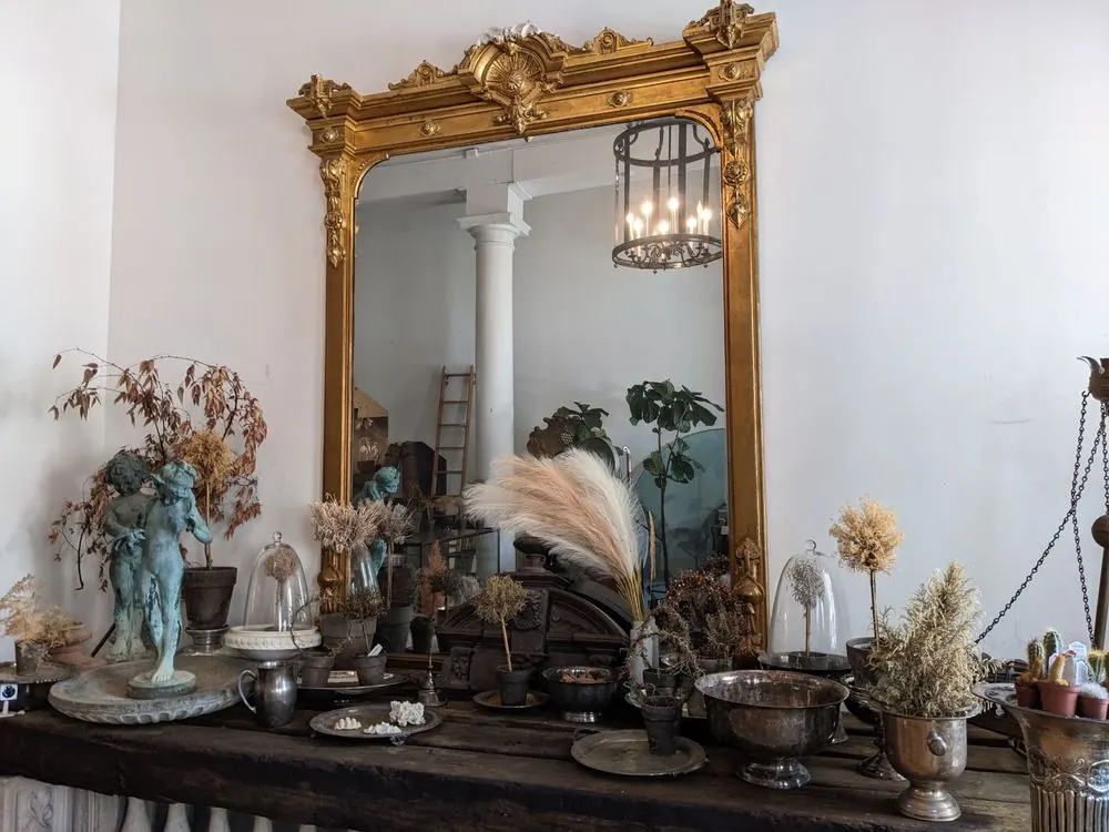 A tall gilt mirror above a table of statues, dried flowers and silver