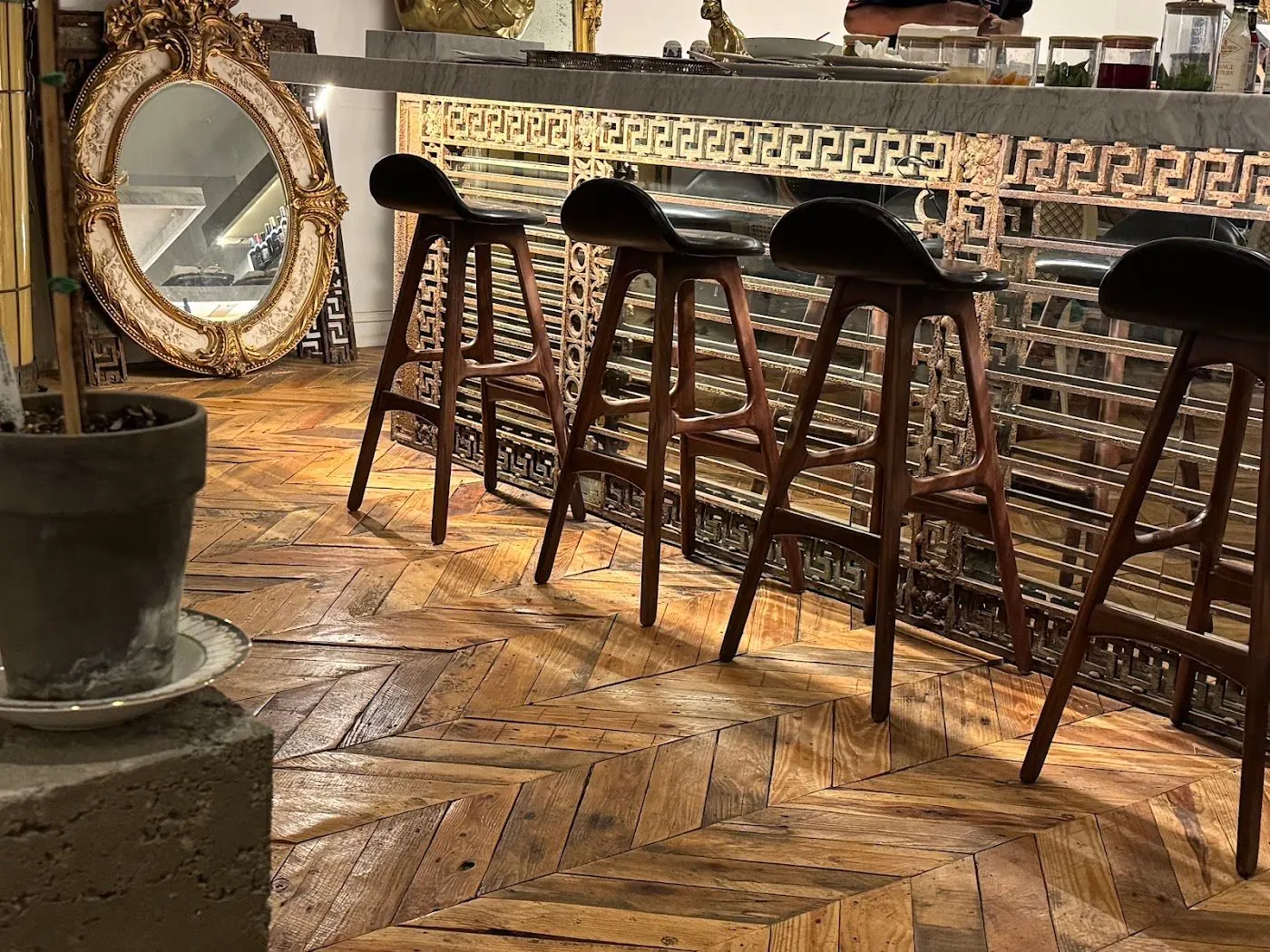 Wooden bar stools before a gilt Greek-key bar front on the herringbone floor, beside an oval gilt mirror