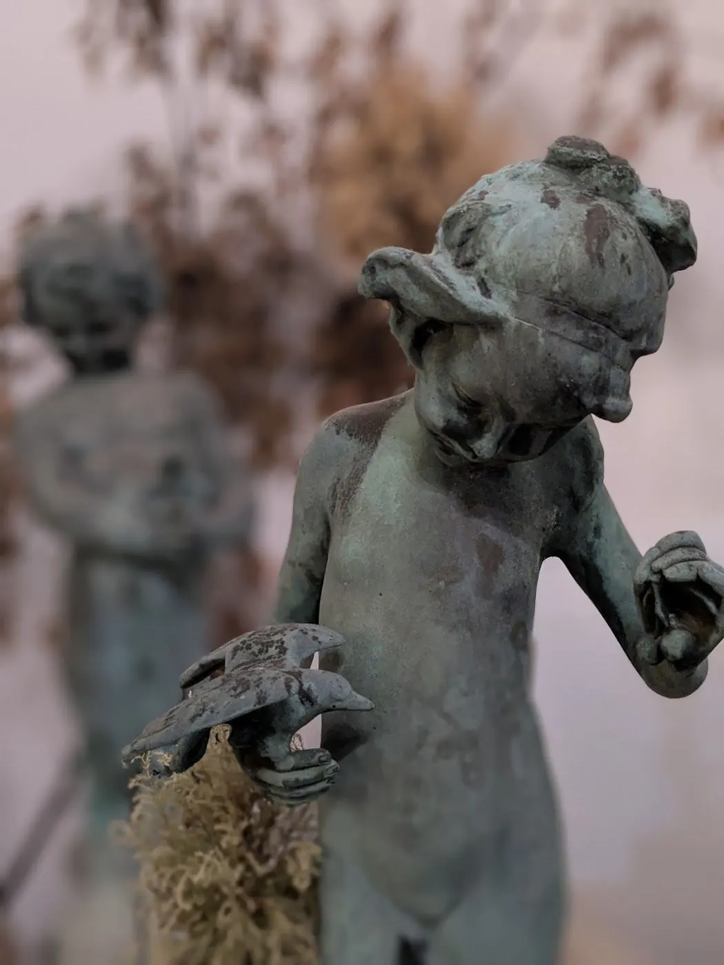 A weathered bronze cherub statue holding a small bird, with dried flowers behind it