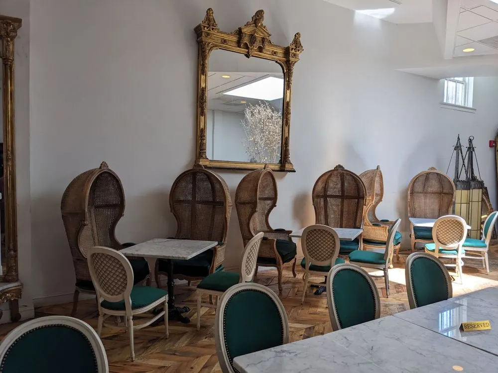 A row of tall wicker porter chairs at marble tables under a large gilt mirror