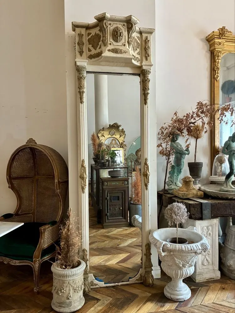 A tall carved white and gold mirror between a porter chair and a stone urn holding a dried topiary